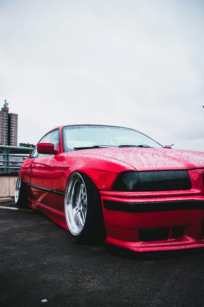 A sleek red car with a lowered stance parked on a wet urban rooftop in a city setting.